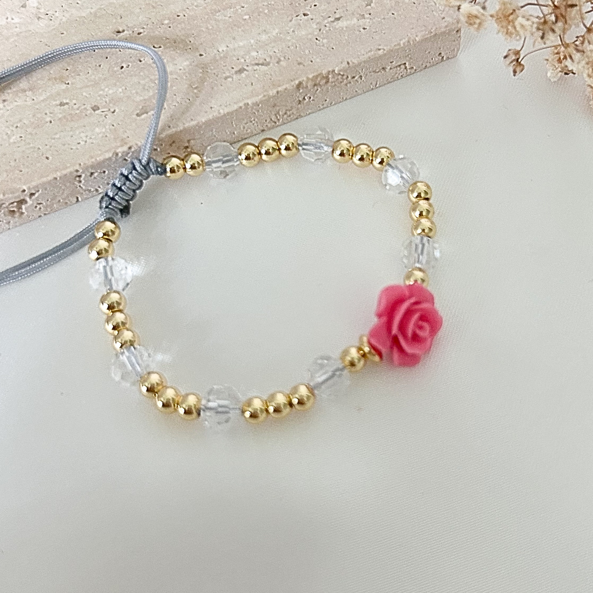 Handmade adjustable bracelet featuring a pink resin rose, sparkling crystal beads, and gold-tone details displayed on a neutral fabric background.