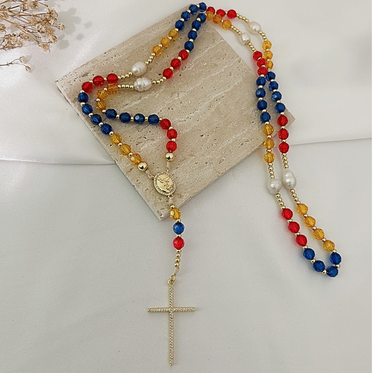 A rosary necklace with red, blue, and yellow faceted beads, white pearl-like beads, a gold-tone medallion, and a textured gold cross, displayed on a beige stone slab over a white fabric background.
