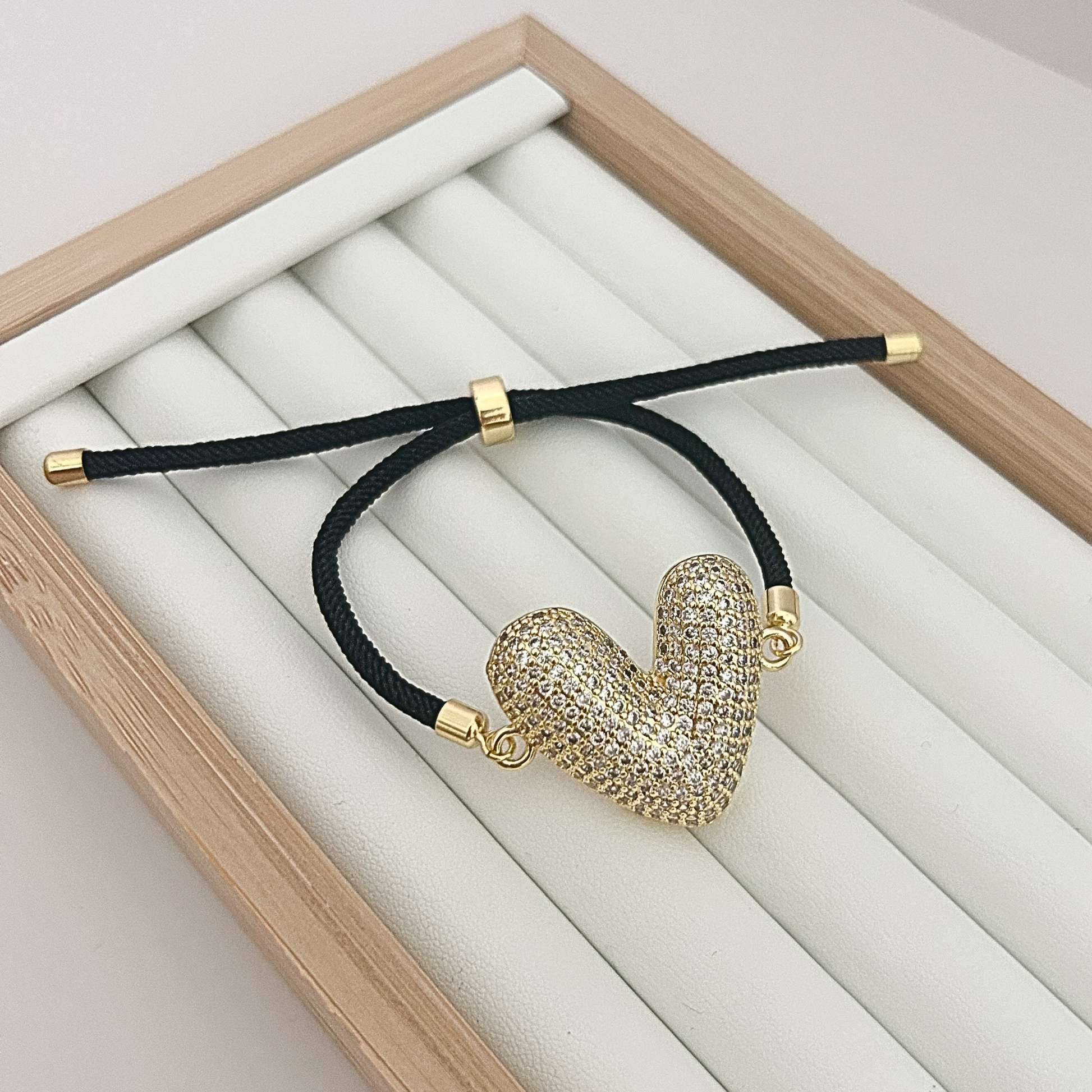 Adjustable black cord bracelet with gold crystal heart charm on jewelry tray.