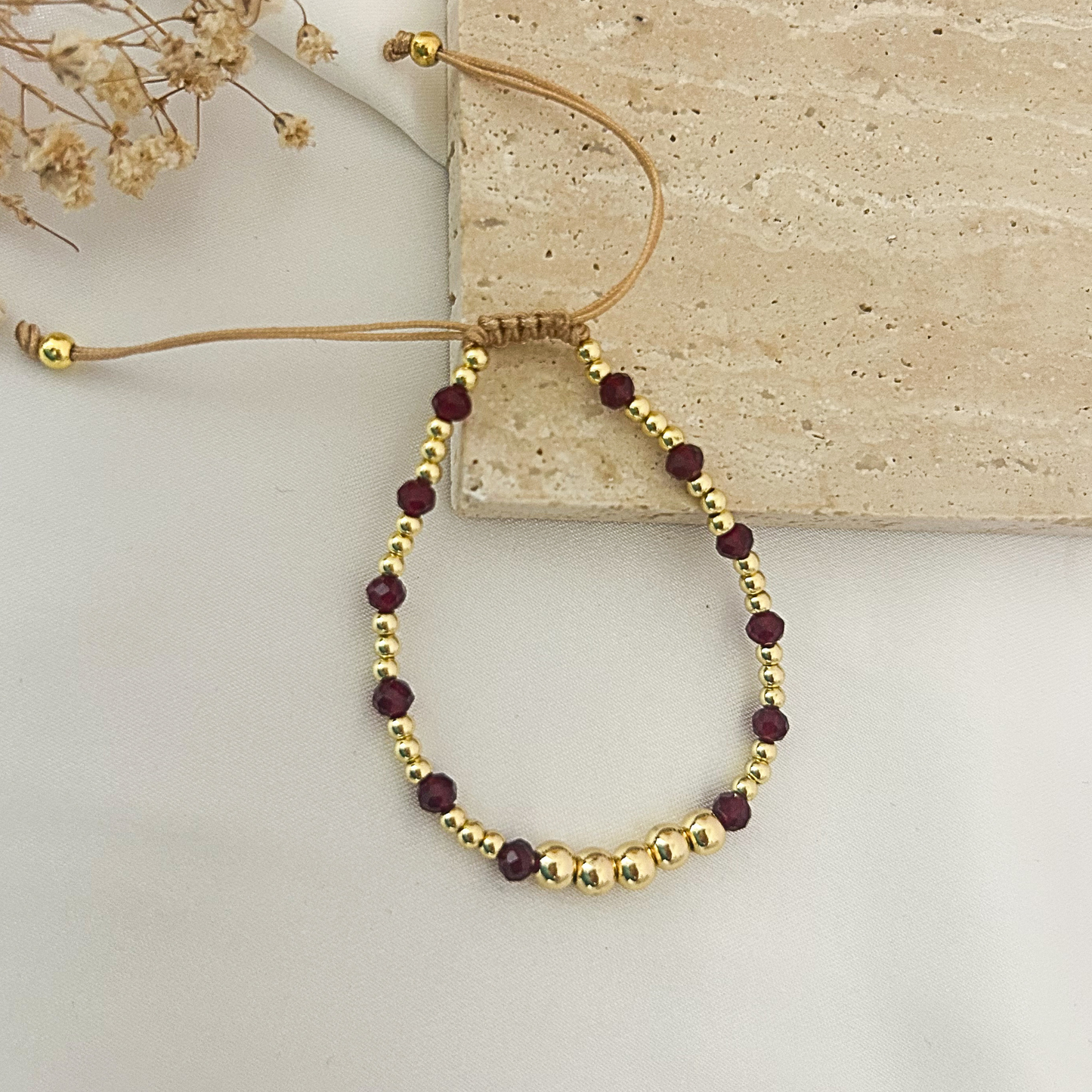 A beaded bracelet with alternating gold-toned round beads and deep burgundy faceted beads, featuring a tan adjustable cord closure, displayed on a light surface with a stone prop.