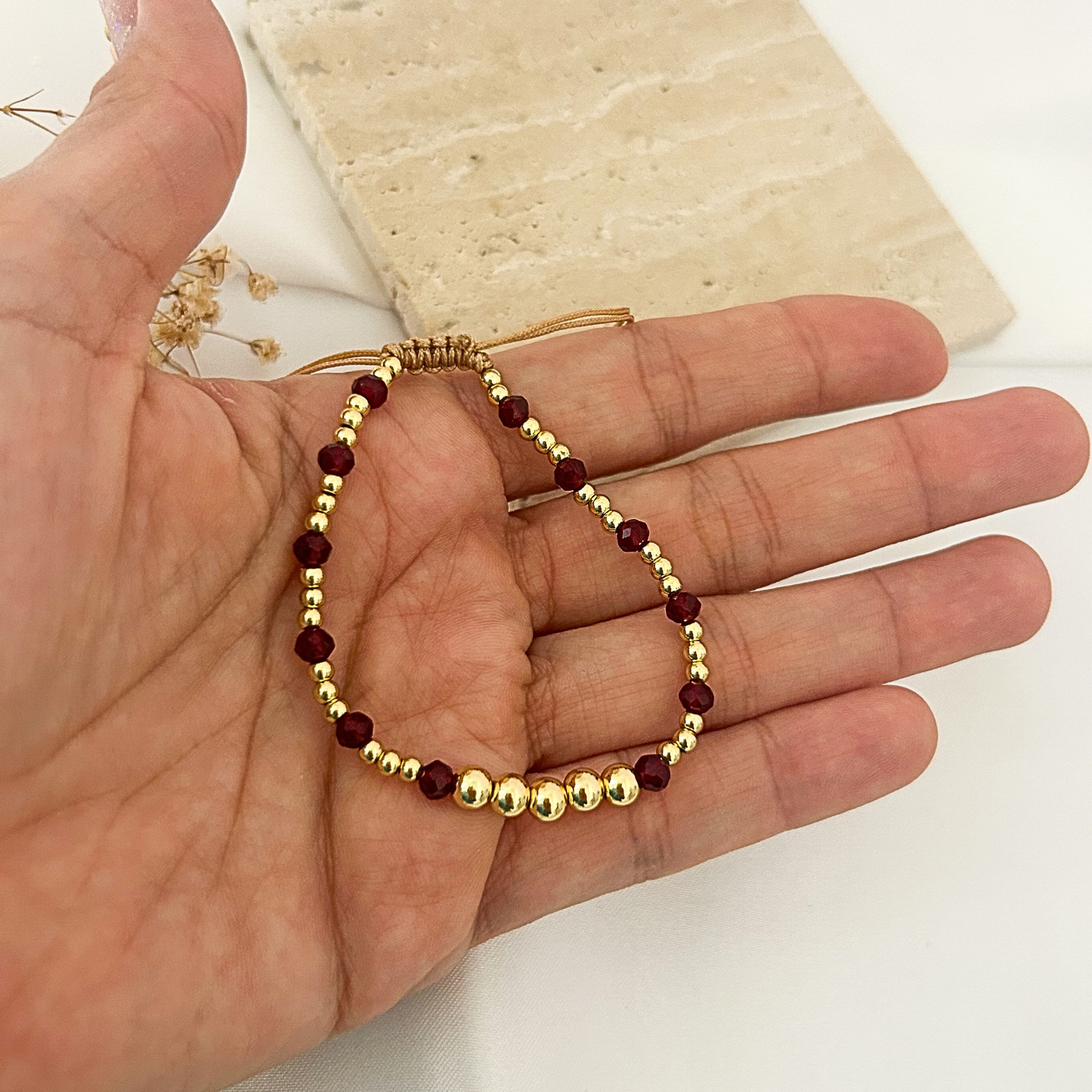 A delicate beaded bracelet with gold-toned round beads and dark red faceted beads, displayed on a person's hand.