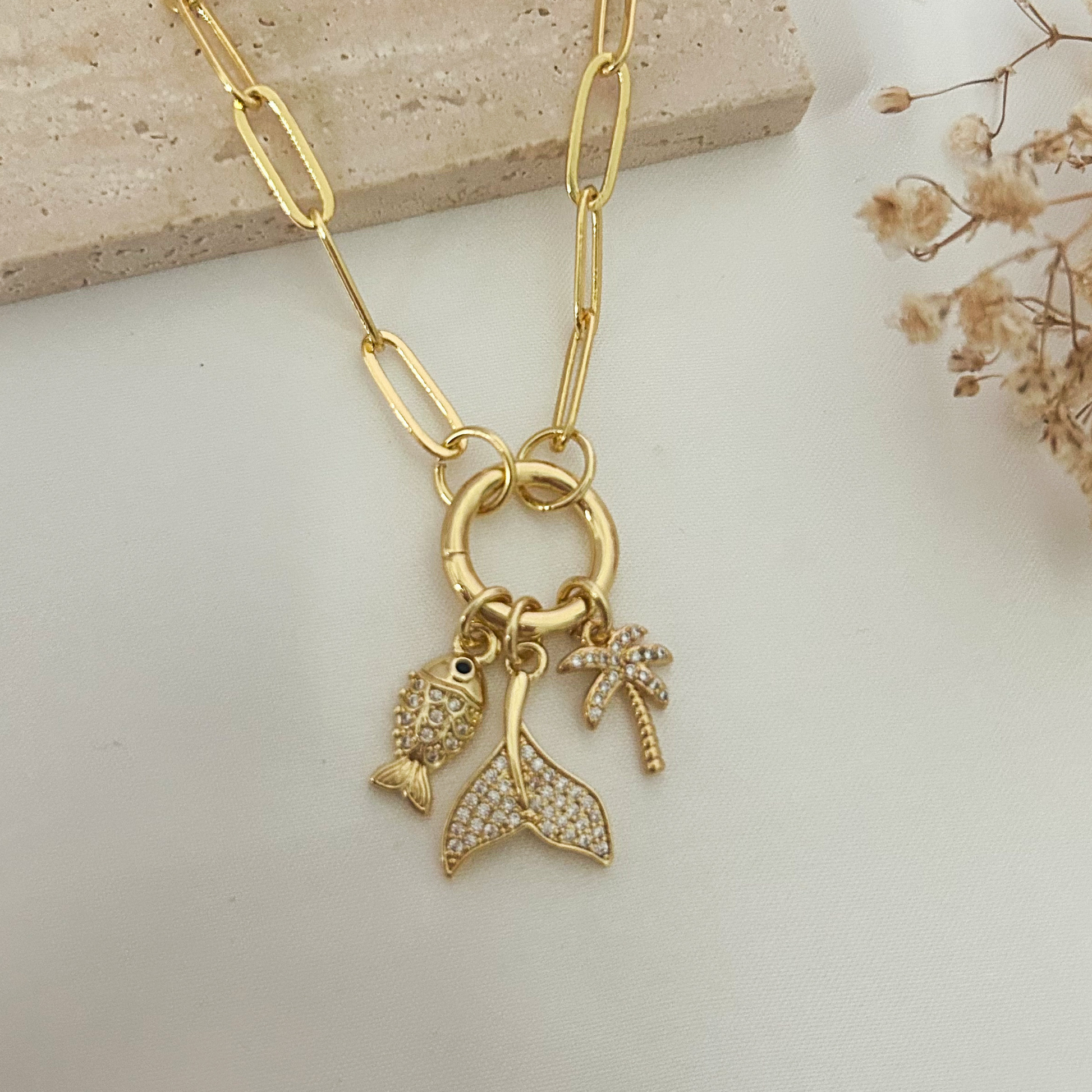 Elegant gold-tone paperclip necklace with tropical charms including a fish, palm tree, and mermaid tail, styled on a soft neutral backdrop for a coastal-inspired look.