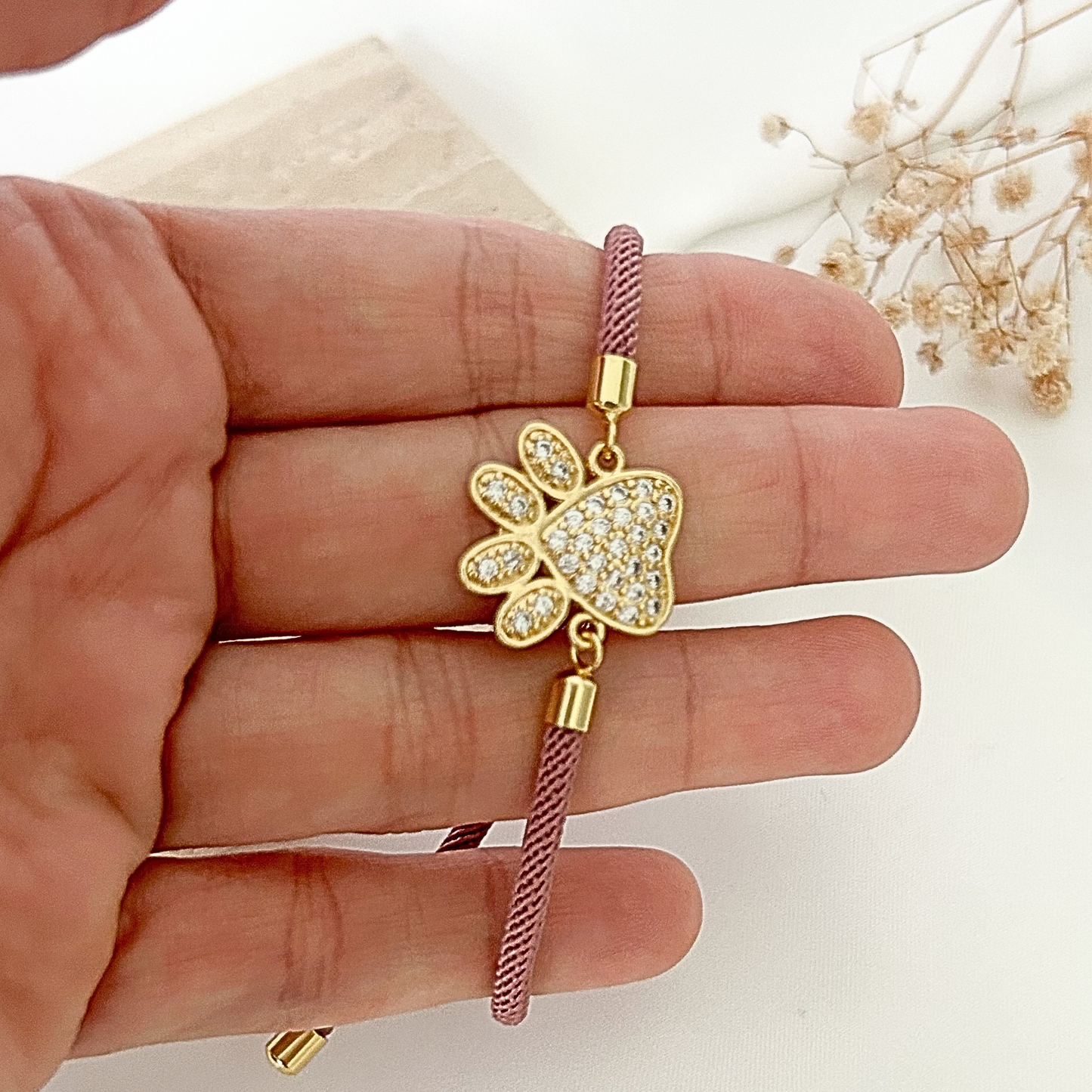 Close-up of a crystal paw charm bracelet held in hand, featuring a dusty mauve braided cord, gold-tone hardware, and sparkling pavé details that catch the light beautifully.