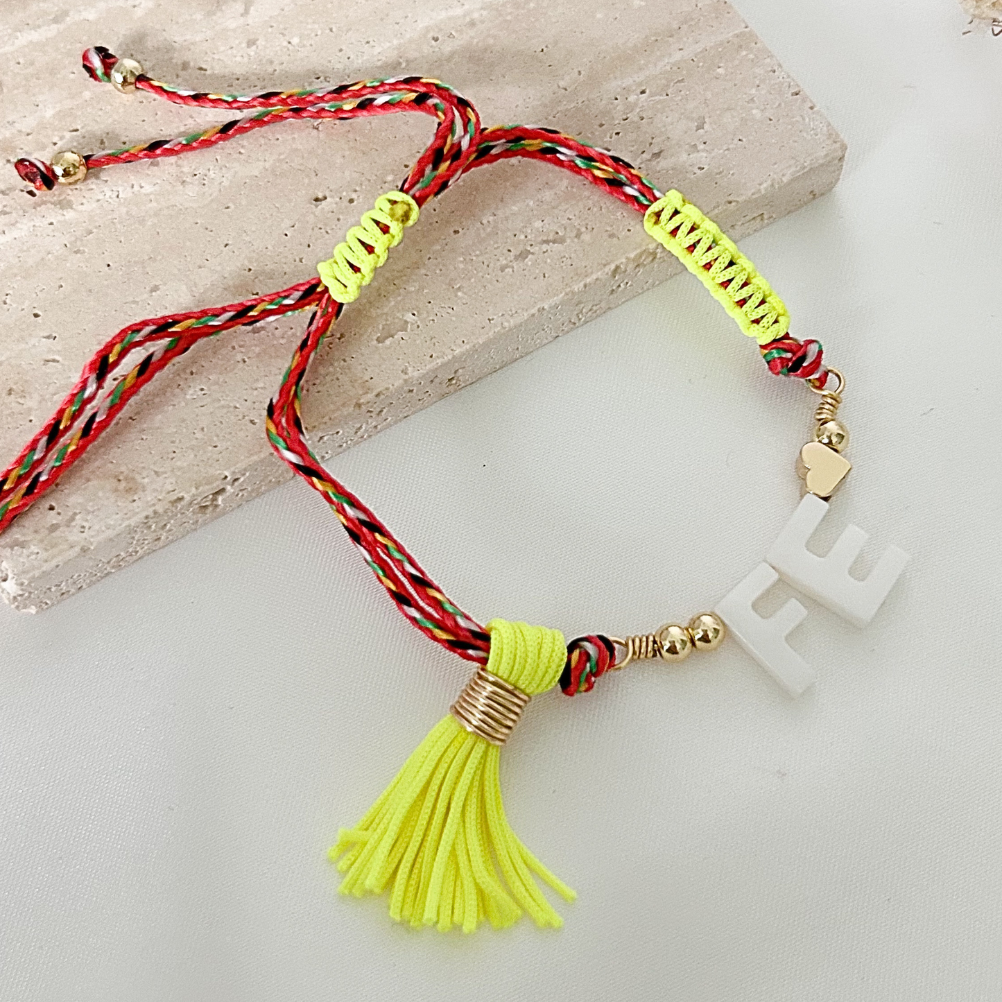 Colorful adjustable bracelet with white “FE” centerpiece, gold-tone details, braided red and green cords, and a neon yellow tassel displayed on a neutral background.