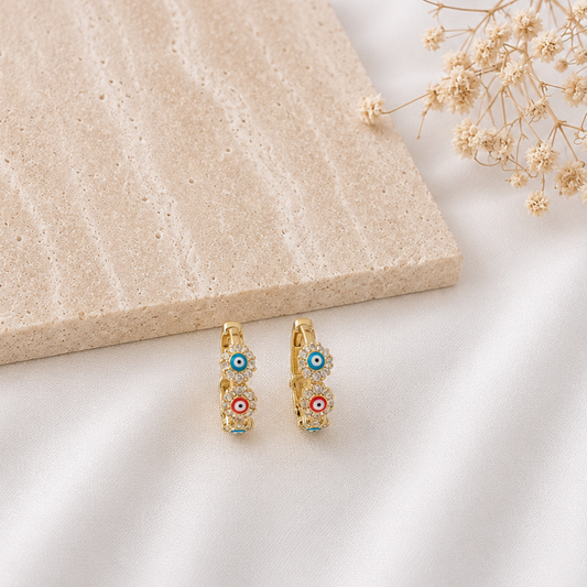 Pair of gold-tone huggie hoop earrings adorned with colorful evil eye enamel charms in blue, red, and turquoise, accented by sparkling cubic zirconia. Stylish protective earrings with a vibrant, modern design, perfect for everyday wear or stacking with other gold jewelry.
