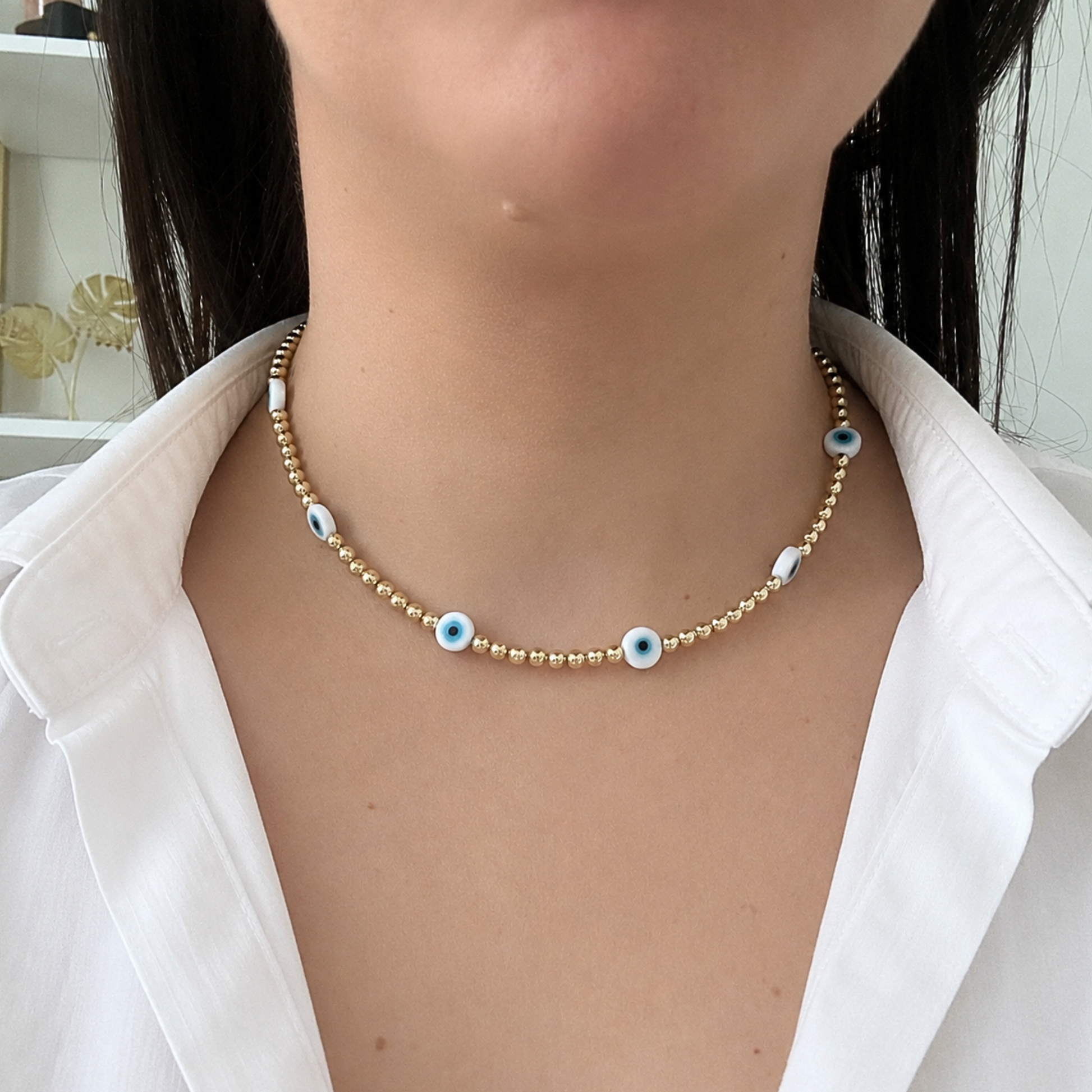 Minimalist gold beaded evil eye necklace worn with a white blouse, featuring blue protective eye accents for a chic layered look inspired by protection and positive energy.