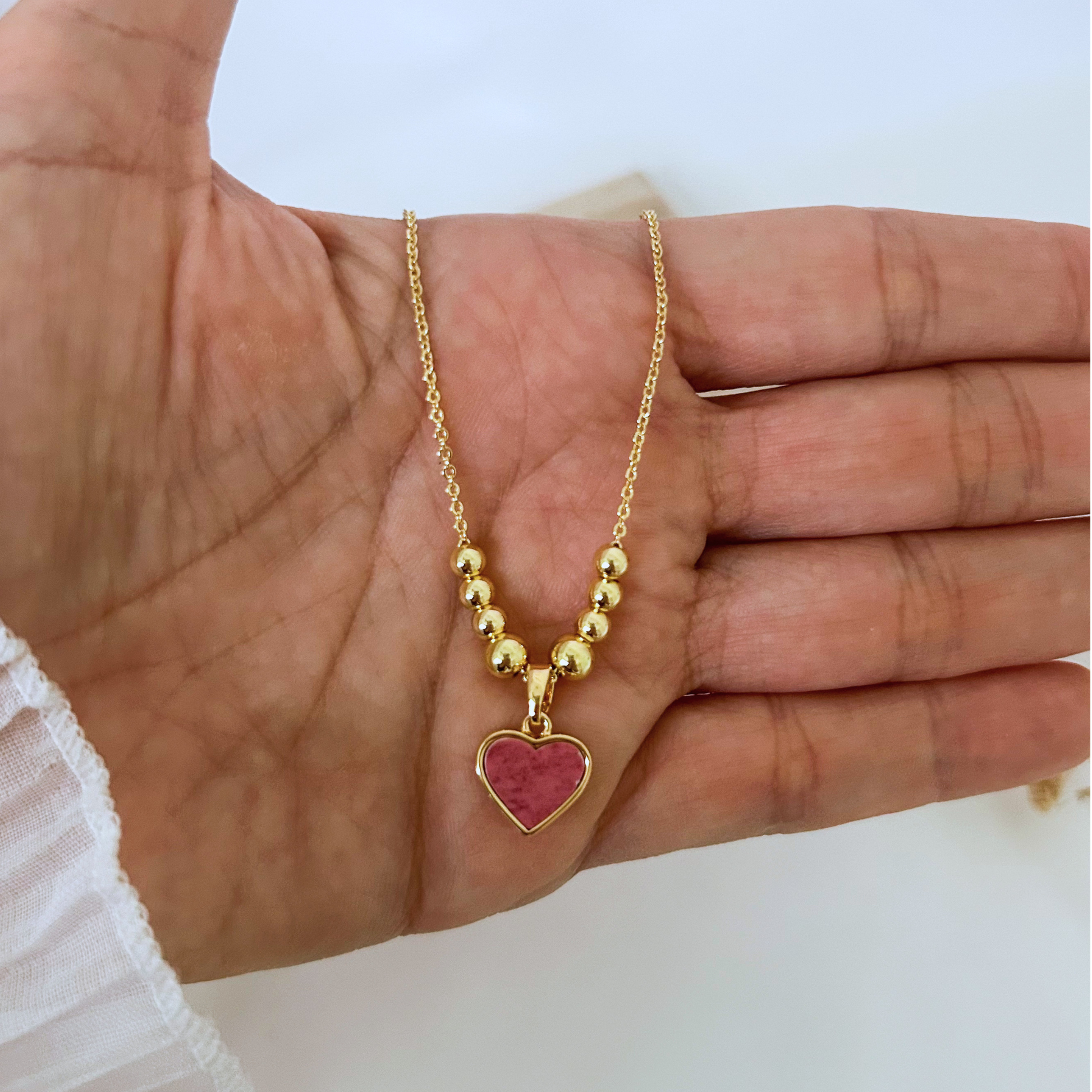 Minimalist gold necklace worn with a white blouse, showcasing a pink heart pendant and gold bead details. A sweet, versatile accessory that adds a subtle pop of color to any outfit.
